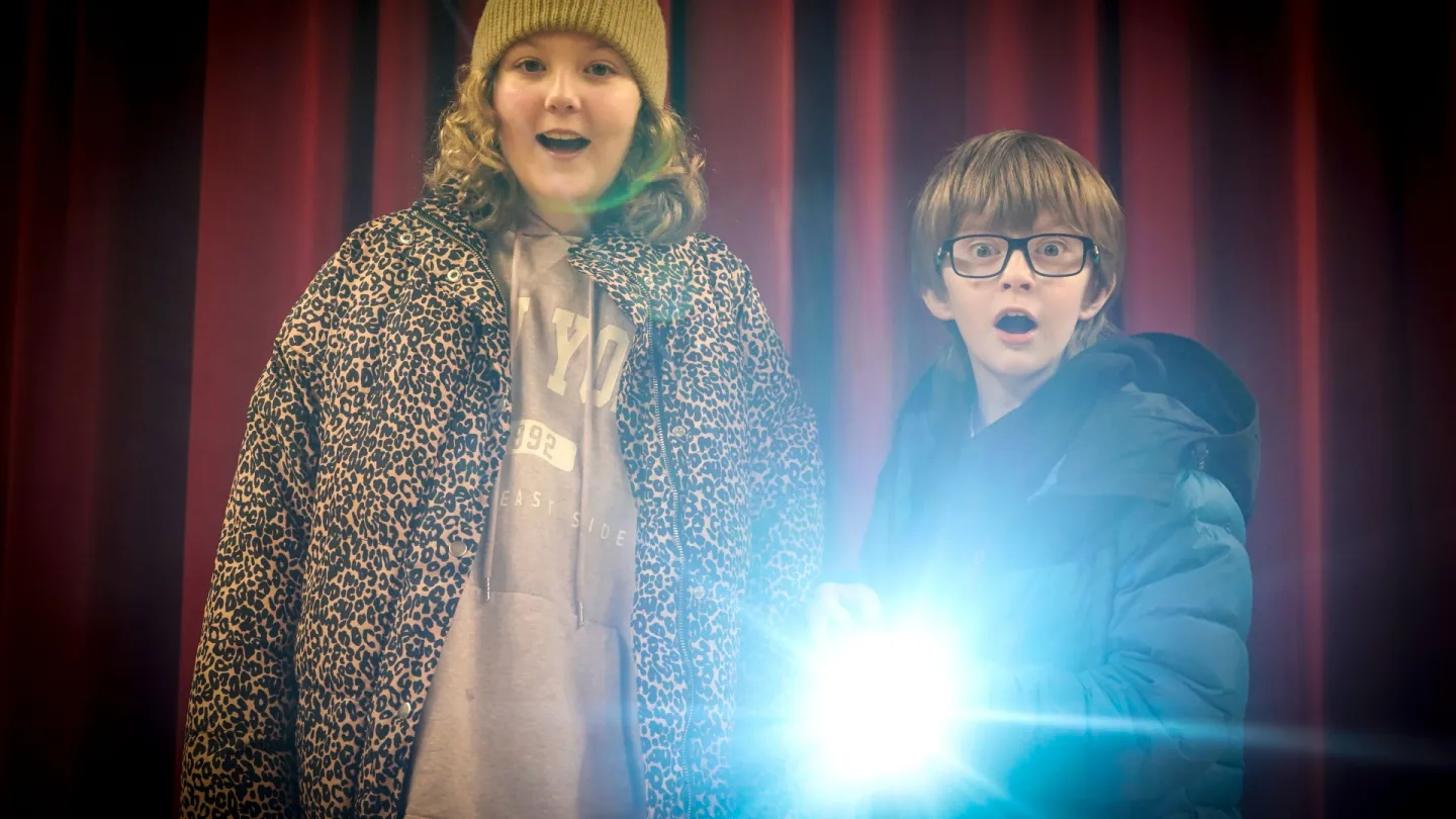 Two people stand before red curtains; one wears a beanie and leopard-print jacket, the other holds a bright light.