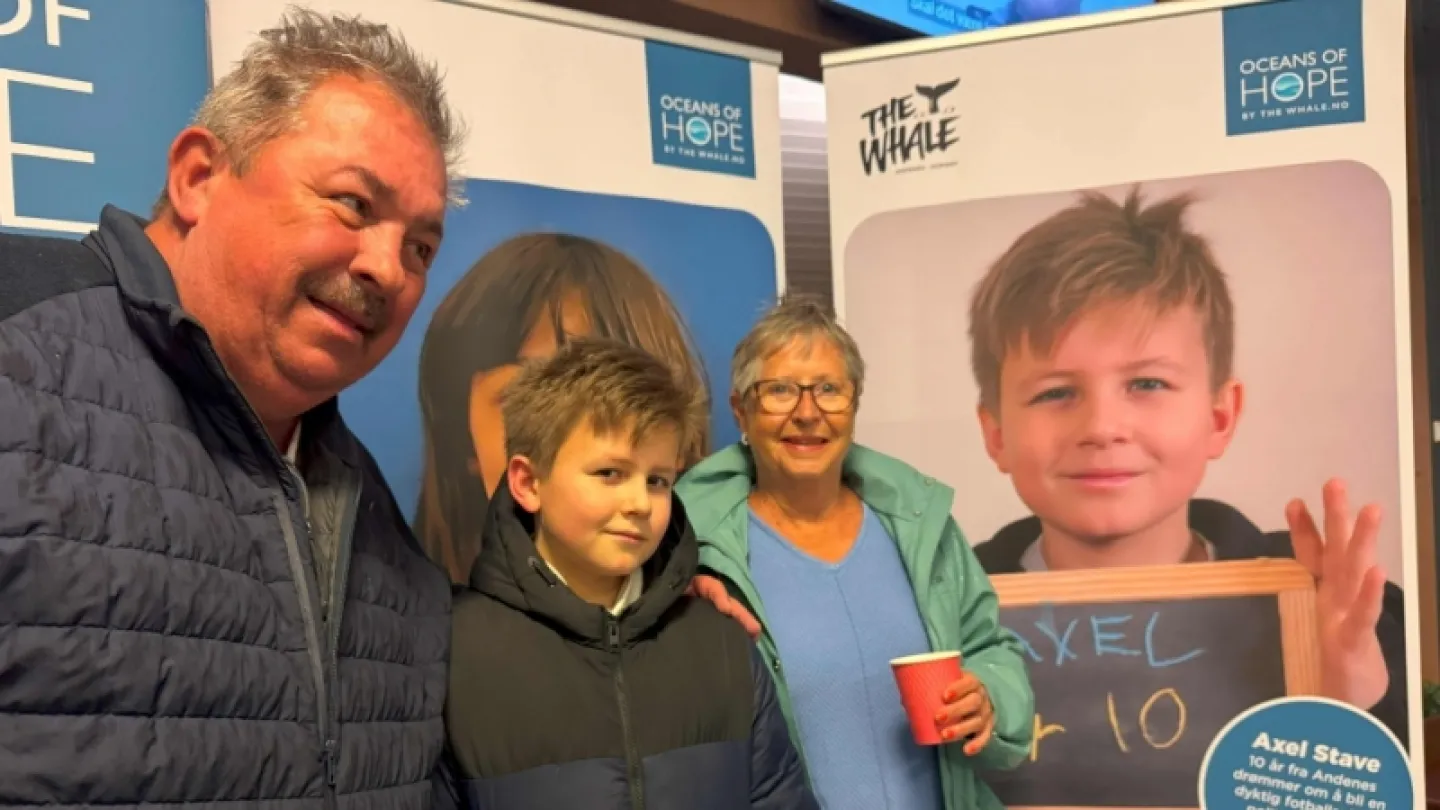 Three people stand indoors in front of blue-and-white informational posters; one holds a red cup.