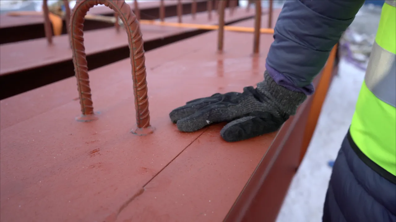 Close-up of a gloved hand resting on a red steel beam with exposed rebar loops at an outdoor construction site.