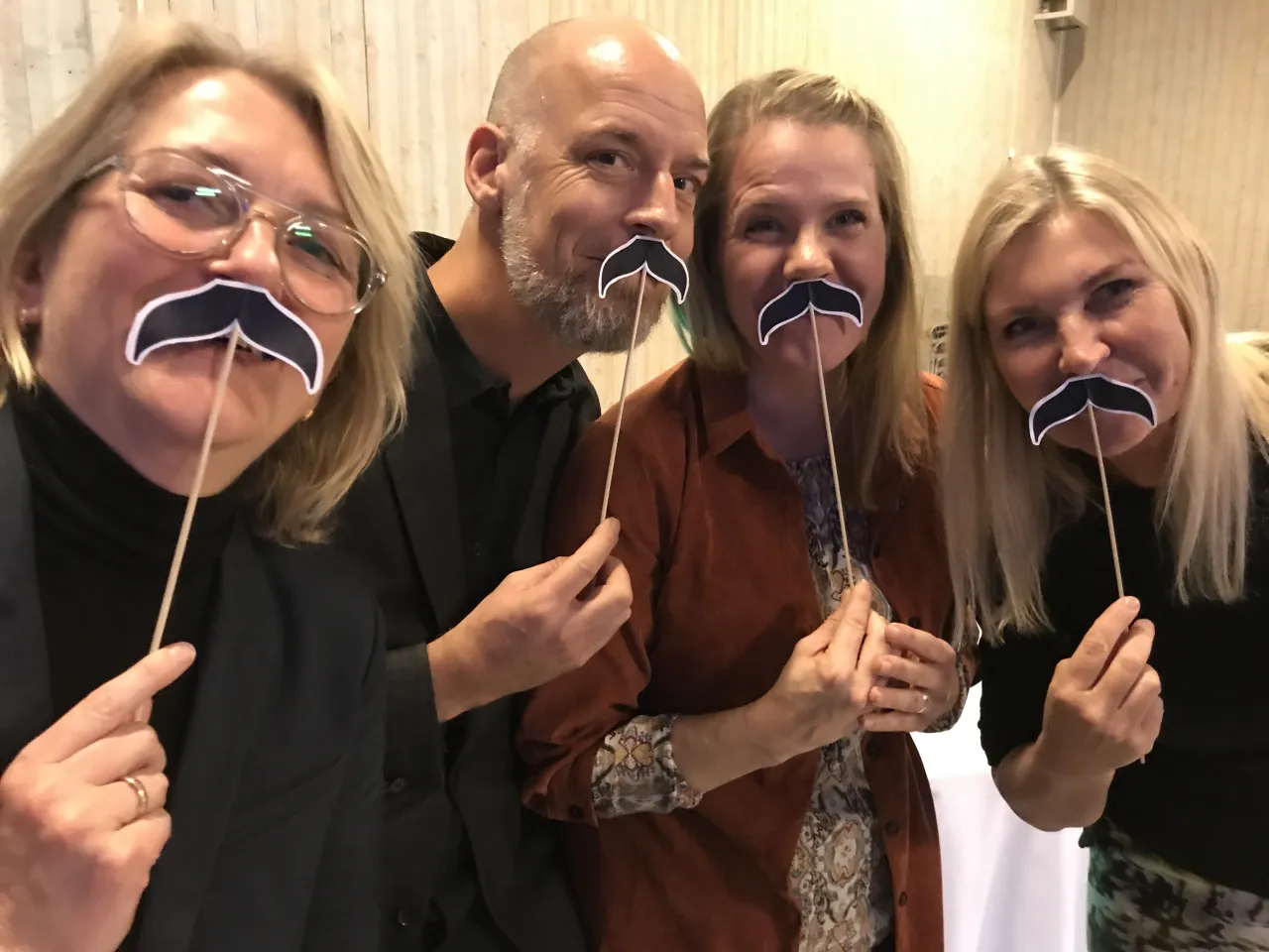 Four adults stand closely indoors, holding stick-mounted rectangular props in front of their heads at an event.