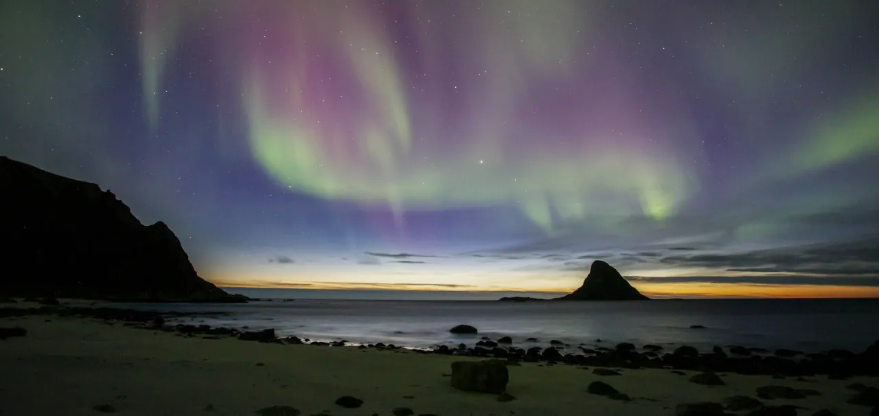 Green and purple aurora streaks above a calm sea at dusk, with a rocky beach and dark island silhouettes.