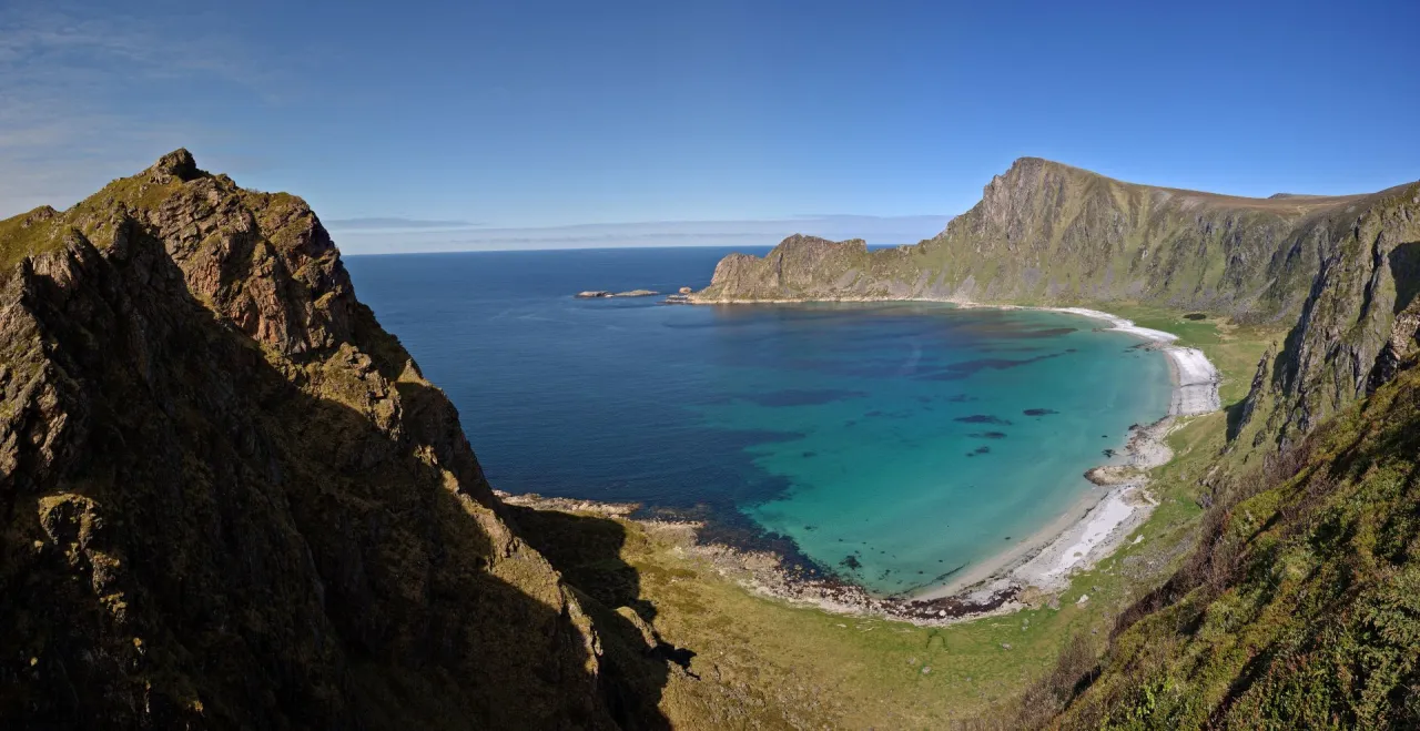 Wide view of a turquoise bay and crescent beach framed by steep rocky mountains under a clear blue sky.