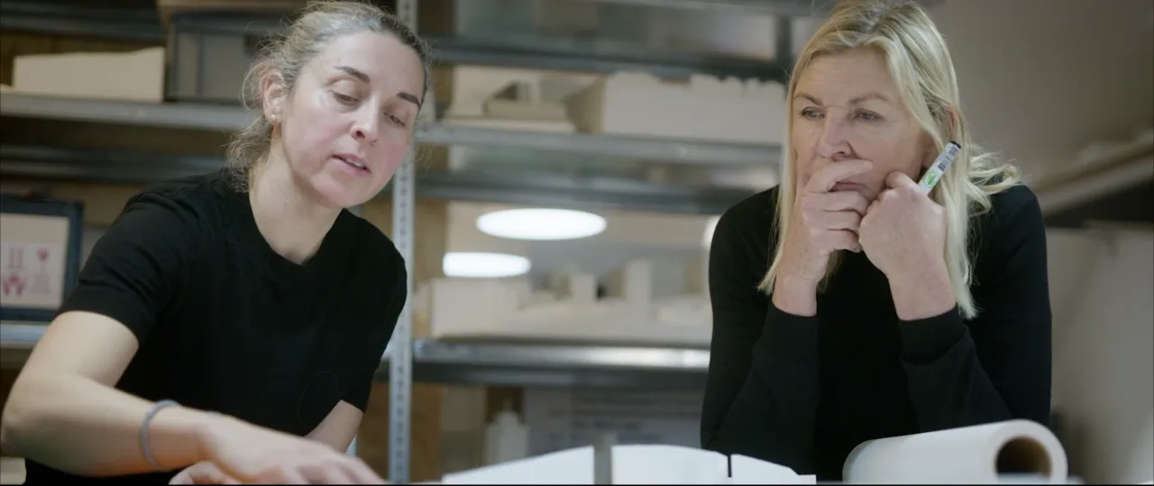 Two people in a workshop studio review papers at a table, with shelves and models in the background.
