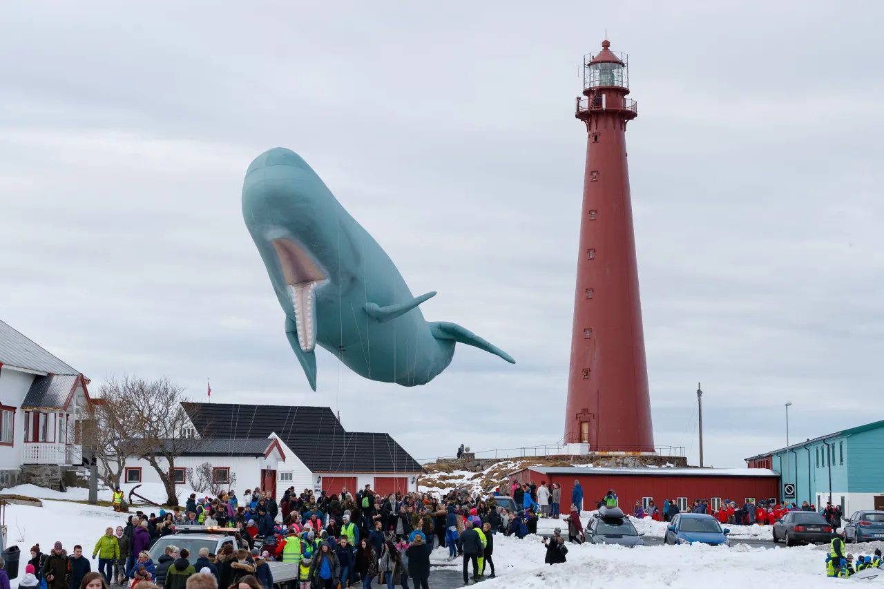 Crowd in snowy coastal village watches a giant blue whale balloon floating near a tall red lighthouse.