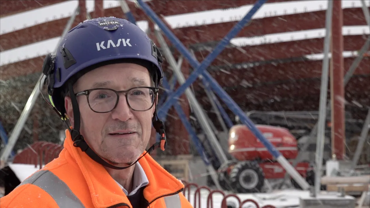Person in an orange high-visibility jacket and blue helmet stands in falling snow at a construction site with scaffolding.