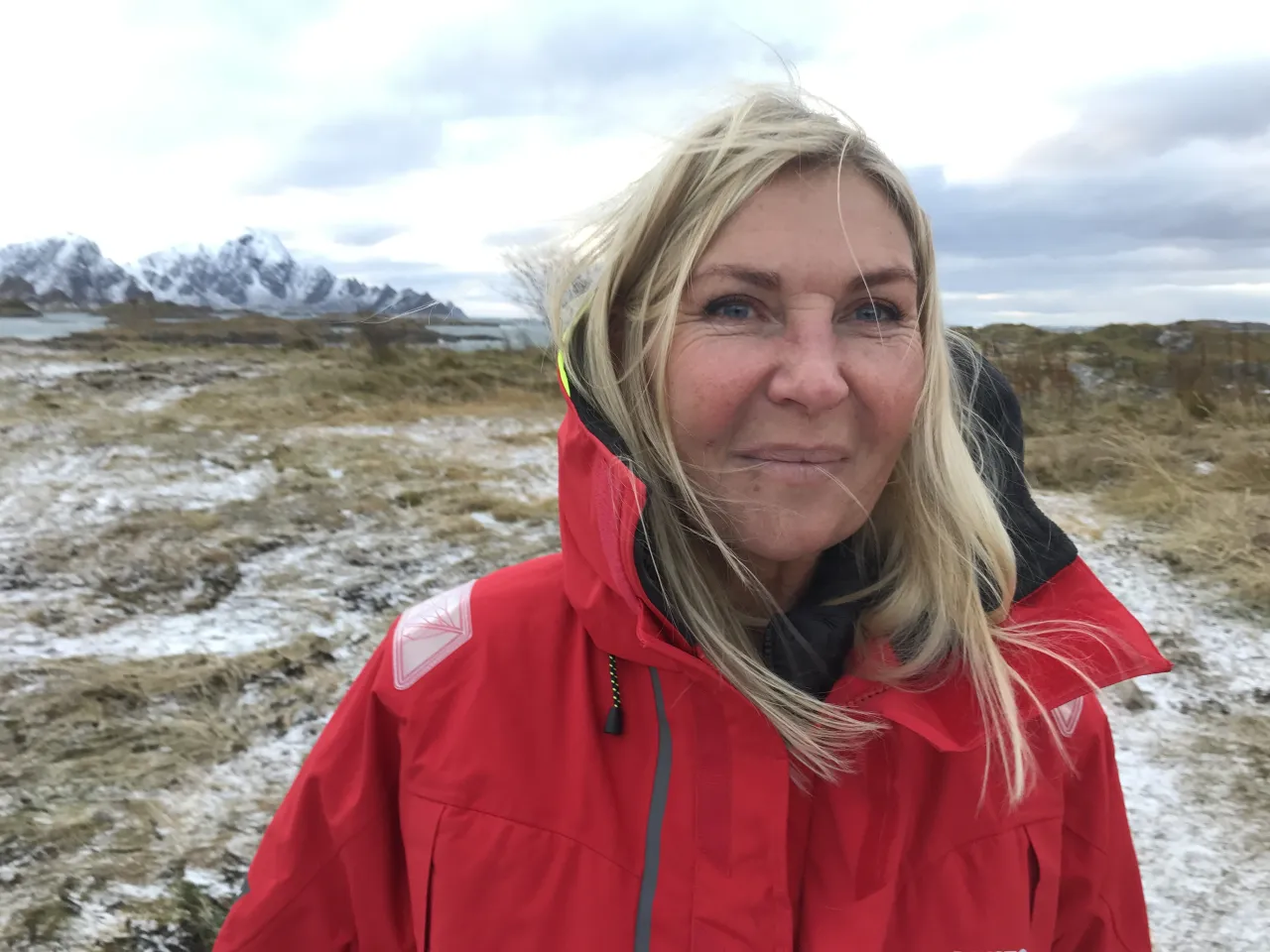 A person in a red hooded jacket stands on a windswept grassy plain with patchy snow and mountains in the distance.