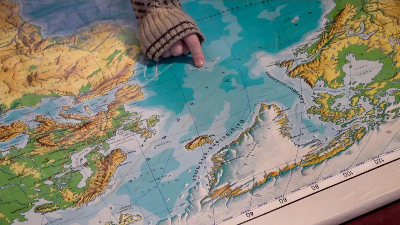 A hand in a knitted sleeve points to a spot in the North Atlantic on a colorful map near Iceland and Greenland.