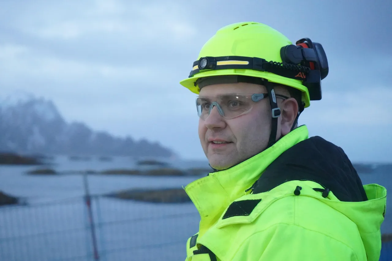 Person in neon safety jacket and hard hat with earmuffs stands near a coastal area under a cloudy sky.