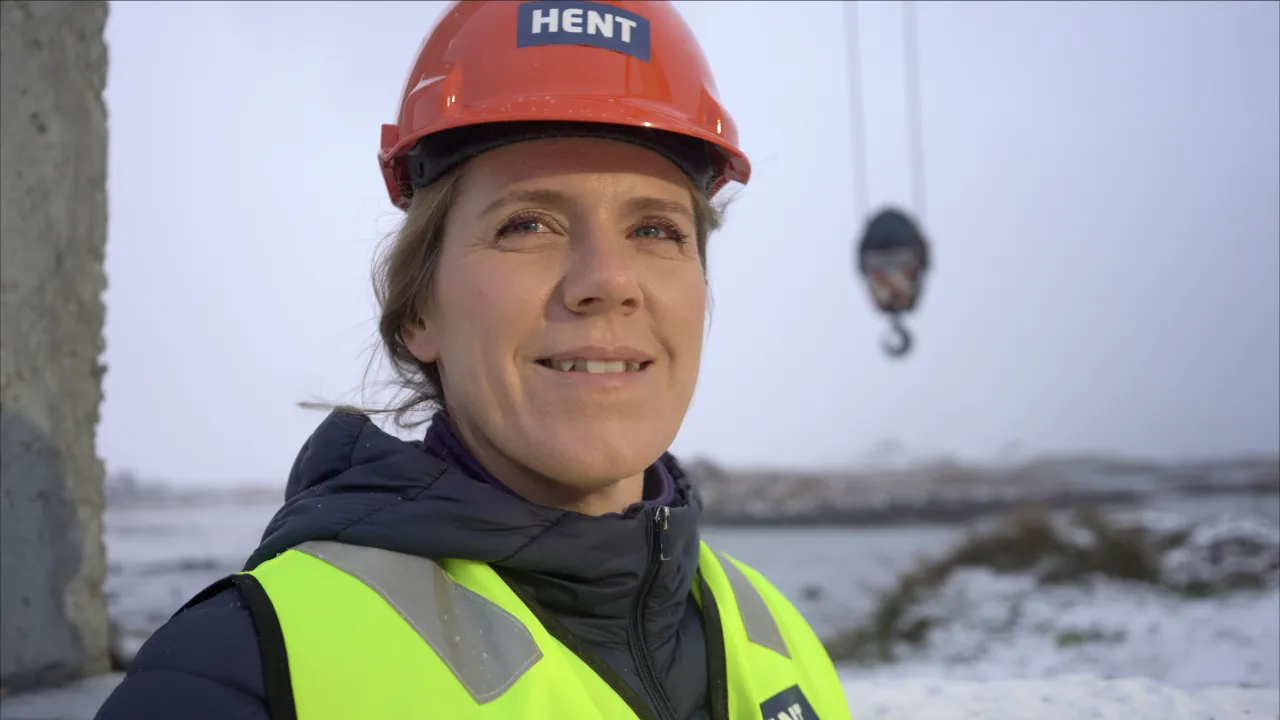Person wearing an orange hard hat, neon safety vest, and winter jacket stands outdoors near a snowy construction area.