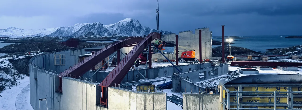 Concrete construction site with steel beams and lift machinery, set by a cold sea with snowy mountains under dark clouds.