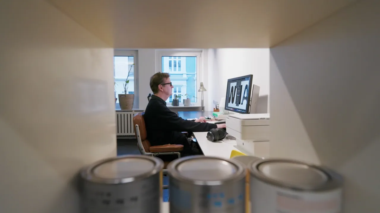 Person seated at a desk in a small office, working on a computer; paint cans in the foreground and windows behind.