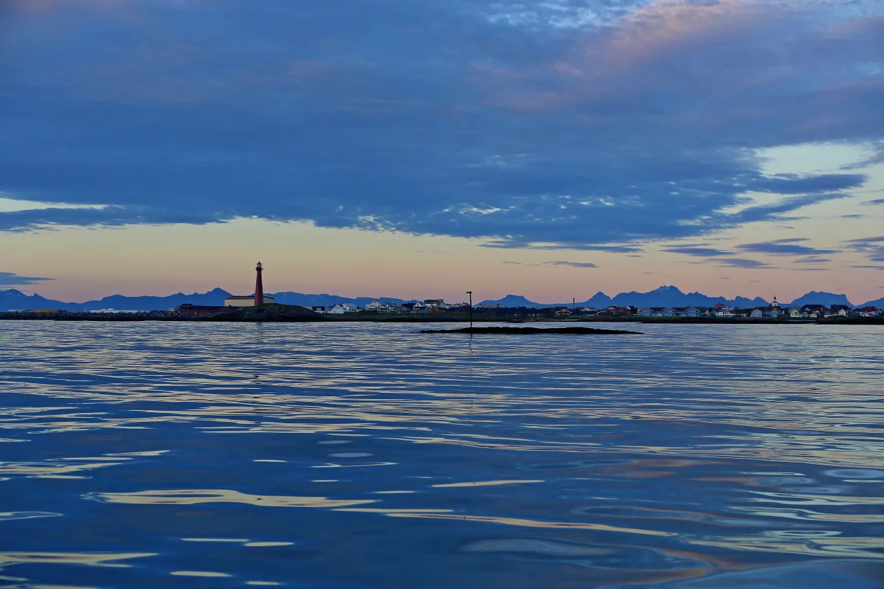 A lighthouse stands on a low rocky shore across calm rippling water, with distant mountains and a cloudy dusk sky.