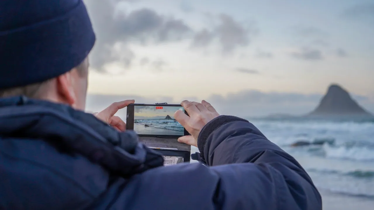 Person in a dark jacket holds a smartphone to record ocean waves, with a rocky island on the horizon at dusk.