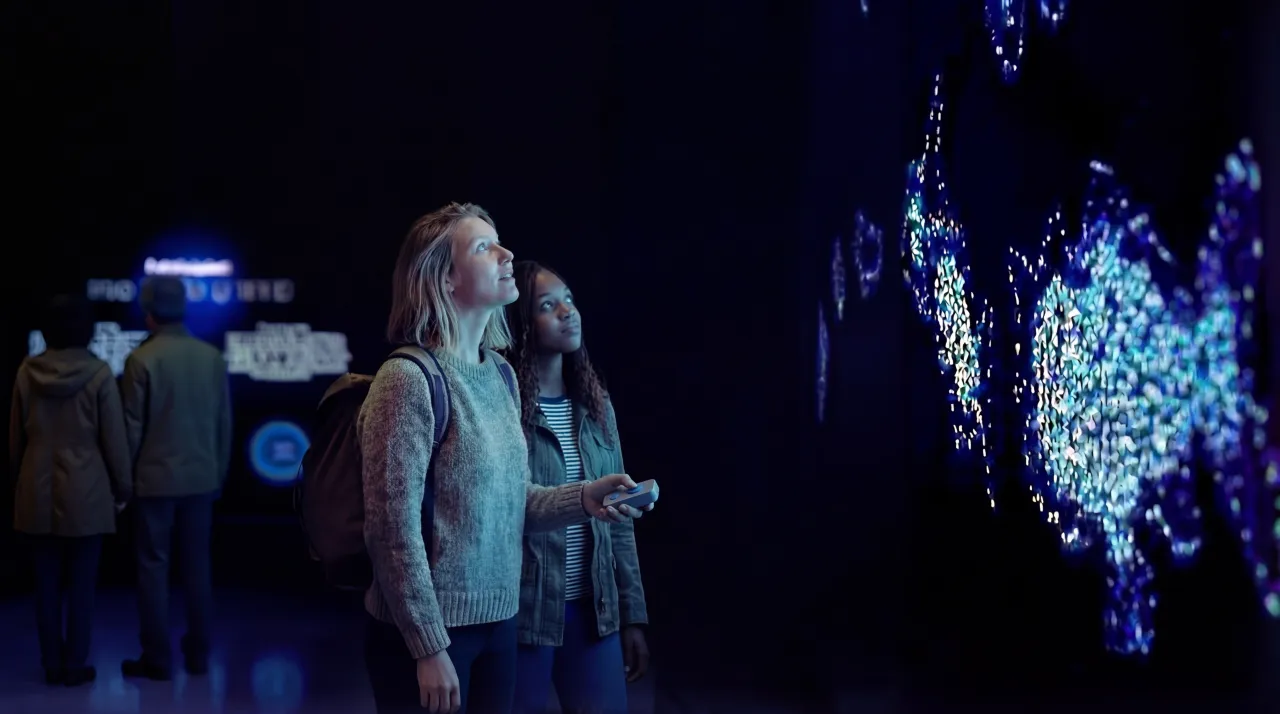 Two visitors stand in a dark exhibition space, viewing a glowing blue interactive light installation on a wall.