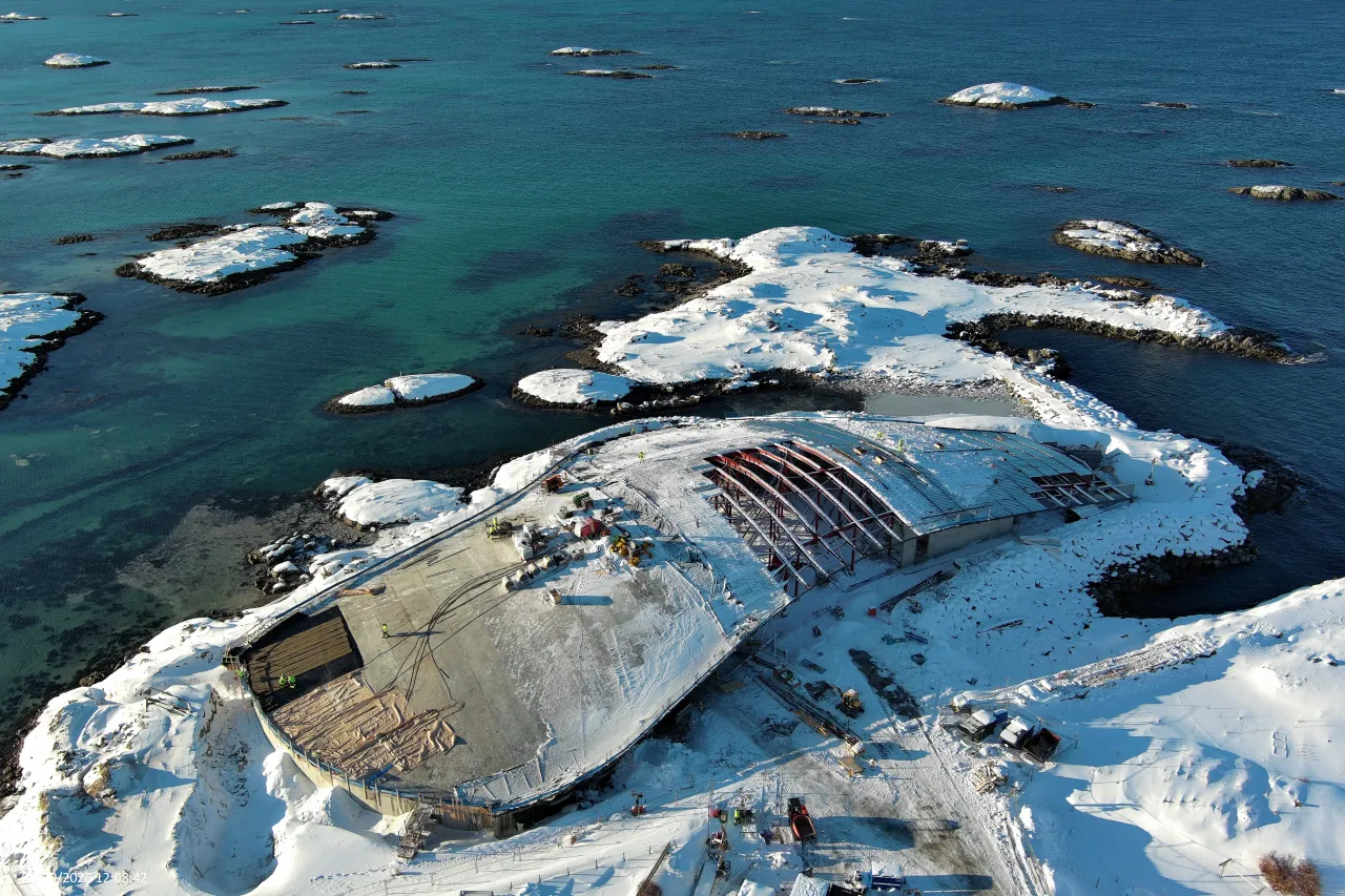 Aerial view of a snow-covered rocky island with a large construction site beside turquoise sea and scattered islets.