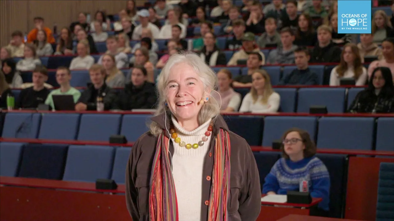 Person standing in front of a filled auditorium with tiered blue seats, wearing a scarf and necklace.