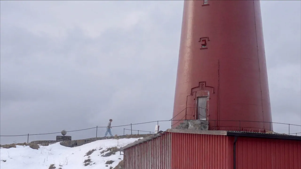 A person walks along a snowy path beside a large red lighthouse tower under an overcast sky.