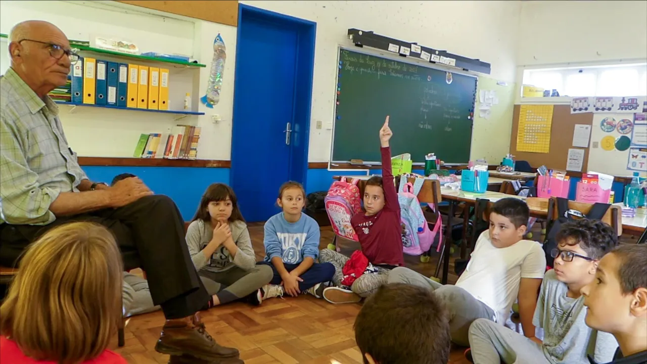 A classroom scene with an adult seated and children sitting on the floor, one child raising a hand near a chalkboard.
