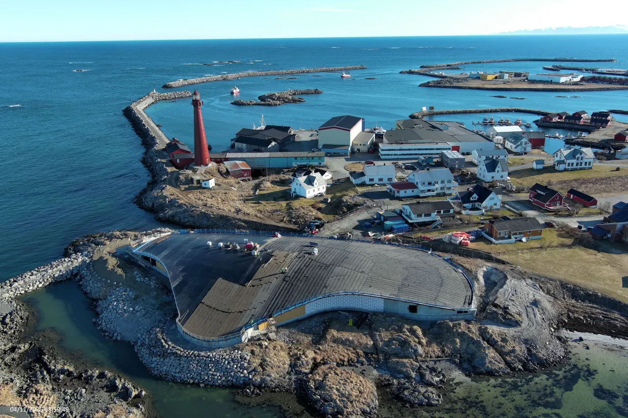 Aerial view of a rocky coastal harbor with a red lighthouse, breakwaters, boats, and buildings beside the sea.