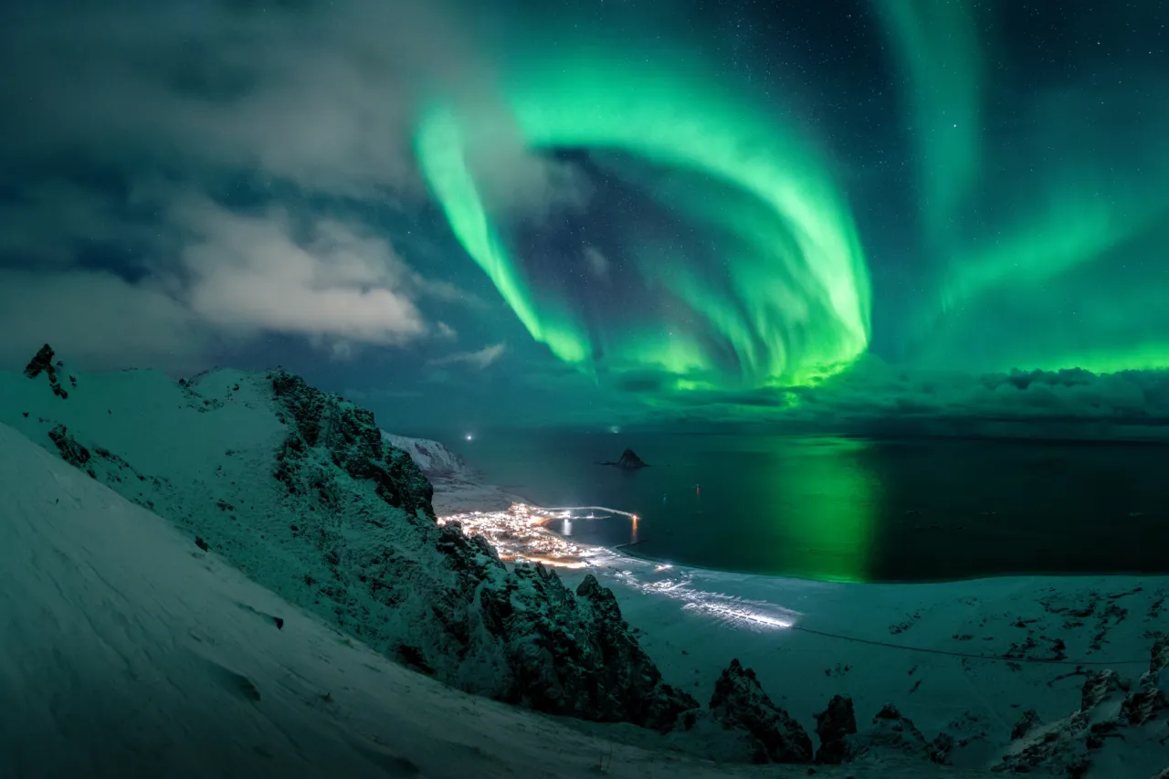 Green aurora lights swirl across a cloudy night sky above snowy mountains and a lit coastal village by the sea.
