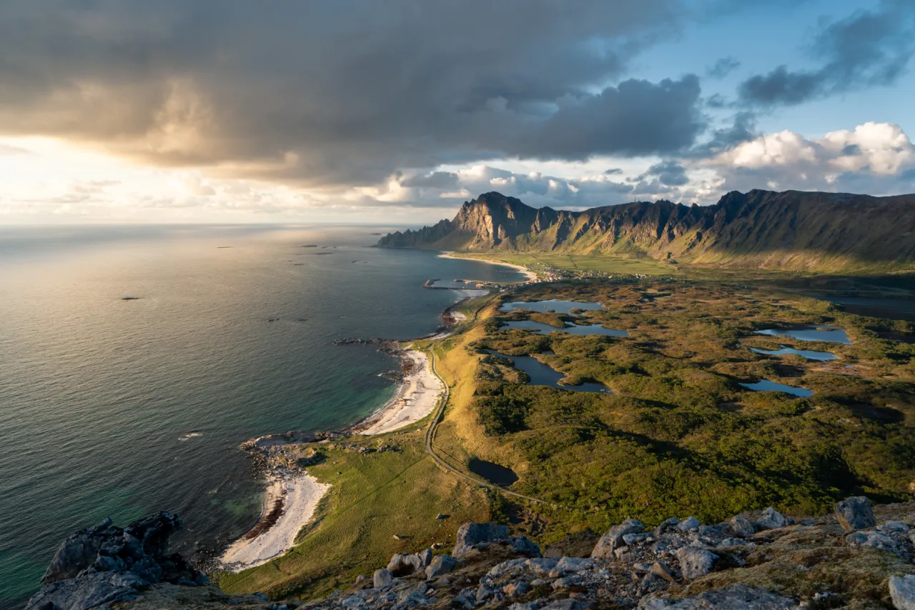Elevated view of a rugged coastline with sandy beaches, green wetlands, and steep mountains beneath dramatic clouds.