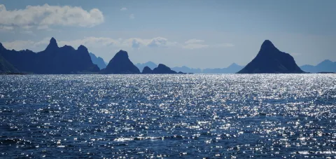 Sparkling blue ocean in the foreground with dark mountain silhouettes on the horizon under a pale sky with clouds.