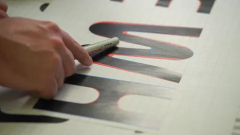 Close-up of a hand using a marker to outline a large black letter on grid paper with red and blue guides.