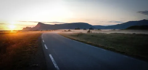 A winding paved road curves through grassy fields with low fog, small houses, and dark mountains at sunrise.