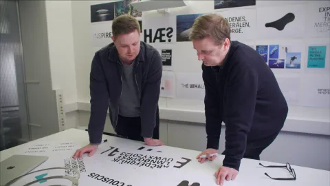 Two people lean over a table reviewing printed design layouts, with posters and photo references on a wall behind.