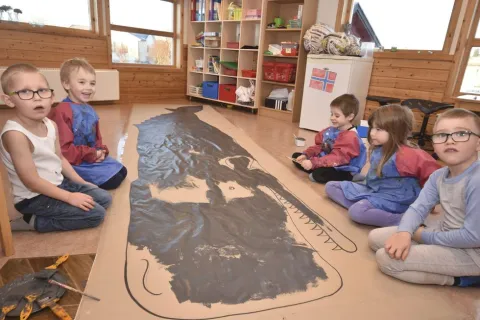 Children sit on a classroom floor around a long paper sheet with a large dark painted animal figure and brushes nearby.