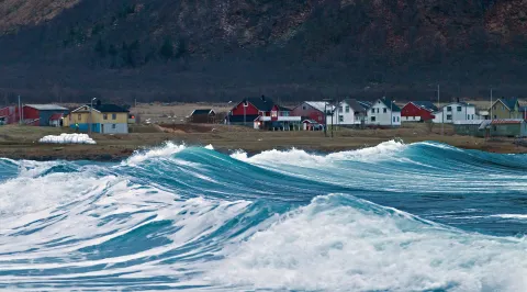 Large blue-green waves crash near a shoreline with colorful houses and barns, backed by a dark hillside.
