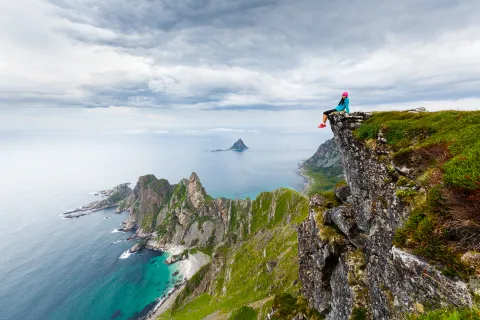 A person in a pink hat sits on a grassy cliff edge above rugged green mountains and turquoise ocean under clouds.