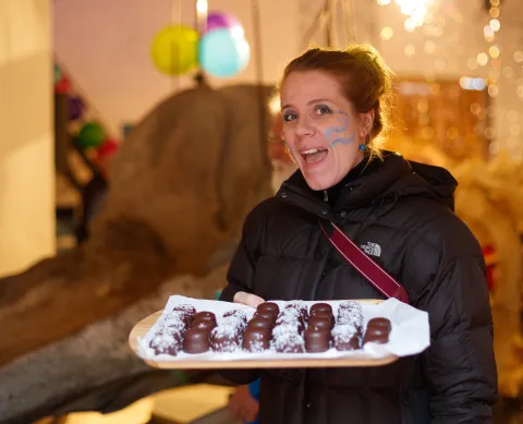 A person in a black puffer jacket holds a wooden tray of chocolate treats indoors with balloons and warm lights behind.