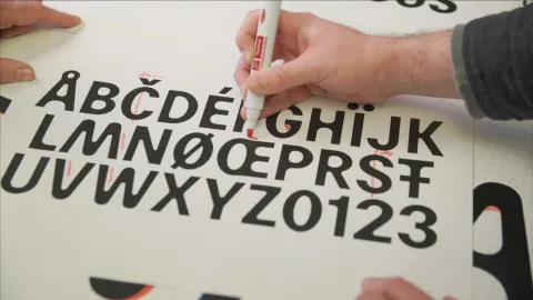 Close-up of hands using a red marker to mark black alphabet letters and numbers on a large printed sheet.