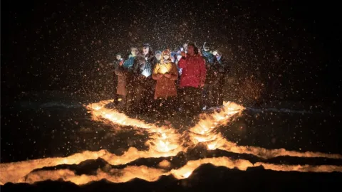 Group in winter coats stands at night behind a winding fire-lit trench, with sparks and smoke in the air.