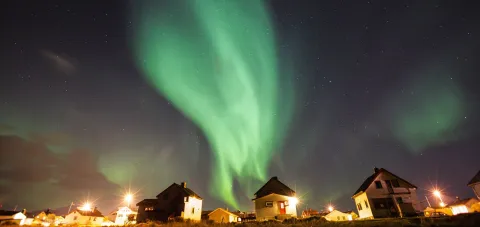 Green aurora borealis swirls across a starry night sky above small houses and streetlights in a village.
