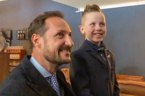 Two men wearing dark coats and ties stand indoors near shelves with bottles and glasses against a blue wall.