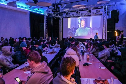 Large audience seated at long tables in a hall, watching a projected speaker on a big screen under blue lighting.