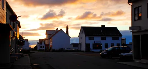Quiet street lined with houses at sunset, with a lighthouse in the distance, parked cars, and orange clouds overhead.