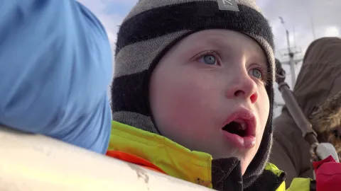 Close-up of a bundled person in a striped knit hat and high-visibility jacket outdoors, with others nearby.