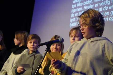 Group of youths on a stage reading from paper sheets beside a microphone, with projected text on a screen behind.