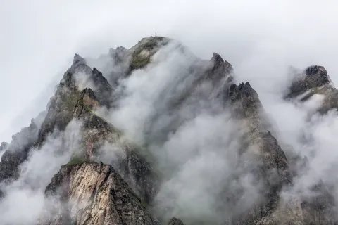 Jagged rocky mountain summit partially covered by thick fog and low clouds, with faint greenery on ridges.