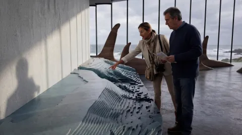 Two adults examine a large layered topographic model in a modern gallery with tall windows overlooking the sea.