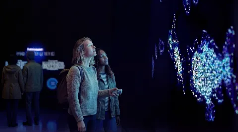 Two visitors stand in a dark exhibition space, viewing a glowing blue interactive light installation on a wall.