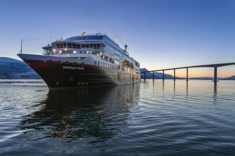 Large cruise ship docked on calm water near a long bridge, with snowy mountains and a blue sunset sky.