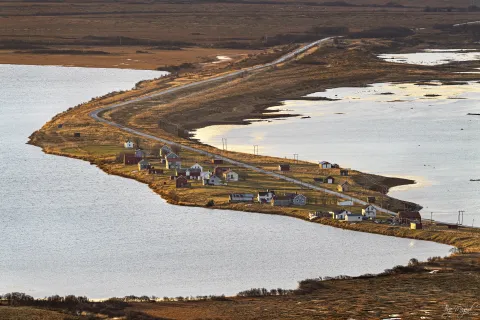 Small cluster of houses along a winding road on a narrow peninsula, bordered by calm water under warm light.