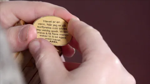 Close-up of hands holding a small round token with printed Norwegian text, against a blurred dark background.