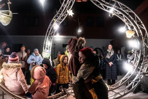 People in winter coats gather outdoors at night beneath a circular metal truss arch with bright lights.