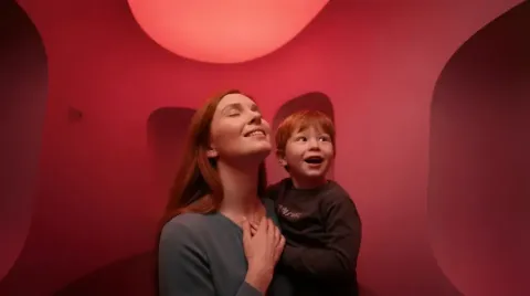 Two people stand close together in a narrow, red-lit interior with curved walls and a large circular ceiling light above.