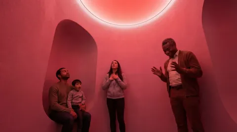 Three adults and a child in a rounded pink room with curved alcoves, gathered beneath a glowing circular ceiling light.